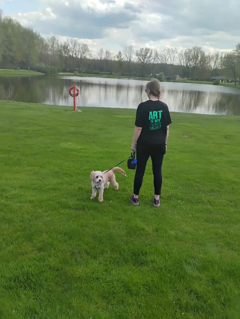 A woman wearing black t-shirt with a tagline "Art is My Favorite Sport" stands near a pond with her dog on a leash.